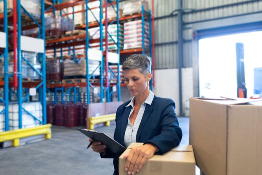Front view of beautiful mature Caucasian female manager checking stocks on clipboard in warehouse. This is a freight transportation and distribution warehouse. Industrial and industrial workers concept