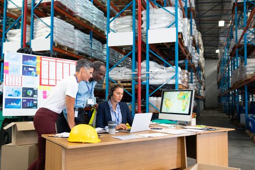 Front view of mature Caucasian female manager with her coworkers discussing over laptop at desk in warehouse. This is a freight transportation and distribution warehouse. Industrial and industrial workers concept