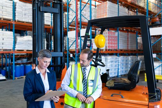 Front view of mature Caucasian male and female staff working together near forklift in warehouse. This is a freight transportation and distribution warehouse. Industrial and industrial workers concept