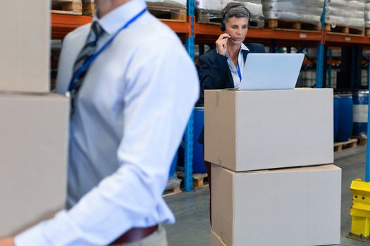 Front view of mature Caucasian female manager talking on headset while working on laptop in warehouse. On the foreground Caucasian male holding cardboard boxes. This is a freight transportation and distribution warehouse. Industrial and industrial workers concept