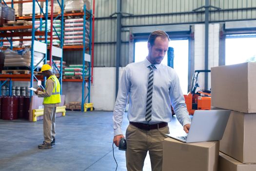 Front view of Caucasian male supervisor working on laptop in warehouse. African-american male worker standing with clipboard in the background. This is a freight transportation and distribution warehouse. Industrial and industrial workers concept