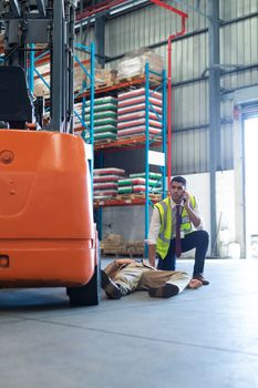 Front view of mixed-race Male supervisor talking on mobile phone while his coworker lying unconscious on the floor in warehouse. This is a freight transportation and distribution warehouse. Industrial and industrial workers concept