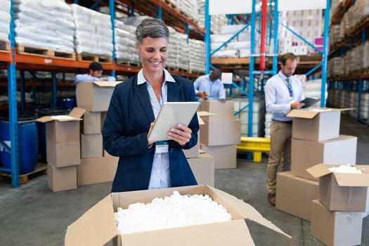 Front view of happy Caucasian female manager working on digital tablet in warehouse. Diverse warehouse workers unpacking behind her. This is a freight transportation and distribution warehouse. Industrial and industrial workers concept