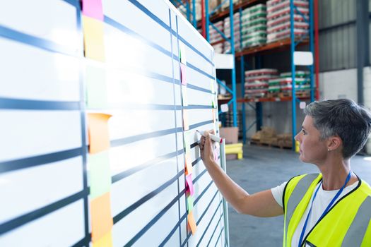 Side view of pretty mature Caucasian female staff writing on whiteboard in warehouse. This is a freight transportation and distribution warehouse. Industrial and industrial workers concept