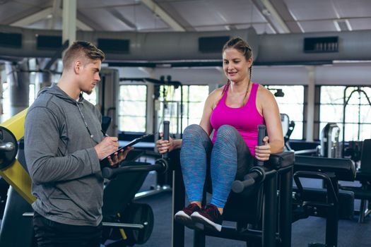 Side view of Caucasian Male trainer assisting young Caucasian female athlete with exercise in fitness studio. Bright modern gym with fit healthy people working out and training