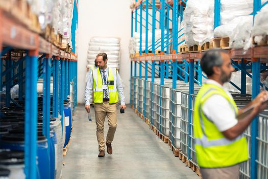 Front view of Caucasian male supervisor walking in aisle in warehouse. In front of him an Asian staff member is writing on clipboard. This is a freight transportation and distribution warehouse. Industrial and industrial workers concept