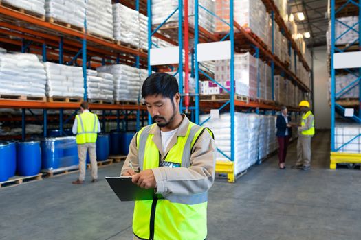Front view of handsome Asian male worker writing on clipboard in warehouse. In the background diverse colleagues working together. This is a freight transportation and distribution warehouse. Industrial and industrial workers concept