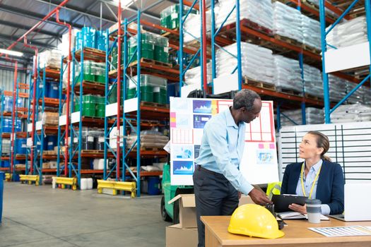 Front view of mature diverse male and female staffs discussing over clipboard at desk in warehouse. This is a freight transportation and distribution warehouse. Industrial and industrial workers concept