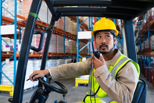 Front view of handsome mature Asian male worker talking on walkie-talkie while driving forklift in warehouse. This is a freight transportation and distribution warehouse. Industrial and industrial workers concept
