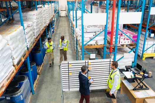 High angle view of mature Caucasian warehouse staff discussing over whiteboard in warehouse. Behind the whiteboard diverse staff are discussing over clipboard in aisle of warehouse. This is a freight transportation and distribution warehouse. Industrial and industrial workers concept