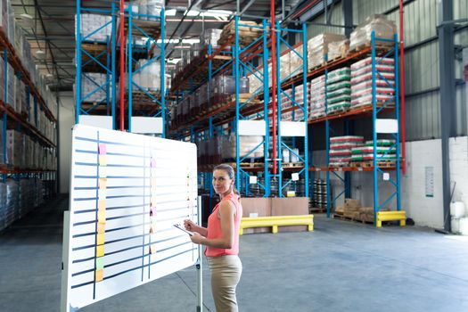Portrait of pretty Caucasian female staff writing on clipboard near whiteboard in warehouse. This is a freight transportation and distribution warehouse. Industrial and industrial workers concept