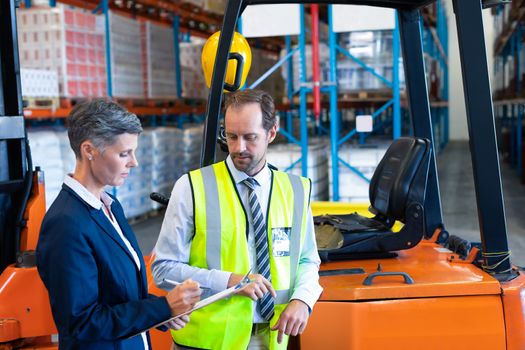 Side view of mature Caucasian male and female staff discussing over clipboard near forklift in warehouse. This is a freight transportation and distribution warehouse. Industrial and industrial workers concept