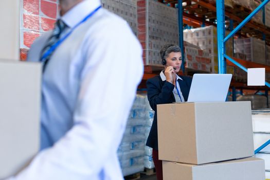 Front view of mature Caucasian female manager talking on headset while working on laptop in warehouse. On the foreground Caucasian male holding cardboard boxes. This is a freight transportation and distribution warehouse. Industrial and industrial workers concept