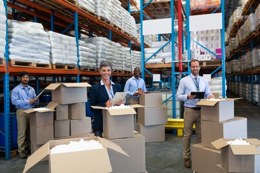 Front view of mature diverse staff looking at camera while working together in warehouse. They are unpacking cardboard boxes. This is a freight transportation and distribution warehouse. Industrial and industrial workers concept