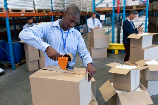Front view of handsome mature African--american male staff packing cardboard box with tape gun dispenser in warehouse. This is a freight transportation and distribution warehouse. Industrial and industrial workers concept