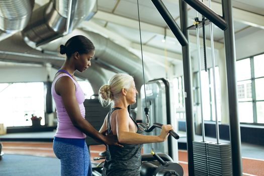 Side view of young fit African-american female trainer assisting active senior Caucasian  woman on lat pulldown machine in fitness studio. Bright modern gym with fit healthy people working out and training