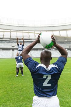 Rear view of African American male rugby player throwing rugby ball in stadium. With players in the background.