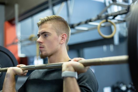 Close-up of handsome fit Caucasian male athletic exercising with barbell in fitness center. Bright modern gym with fit healthy people working out and training