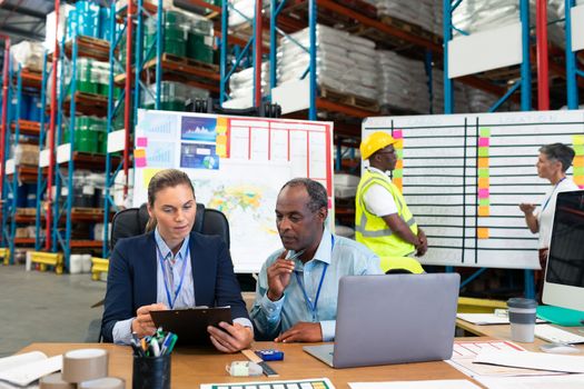 Front view of Caucasian female manager and African-american male supervisor discussing over clipboard at desk in warehouse. This is a freight transportation and distribution warehouse. Industrial and industrial workers concept