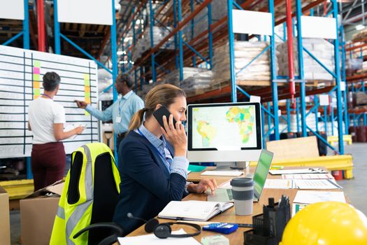 Side view of beautiful mature Caucasian female manager talking on mobile phone at desk in warehouse. In the background diverse coworkers are discussing. This is a freight transportation and distribution warehouse. Industrial and industrial workers concept