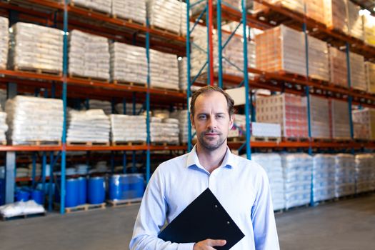 Portrait of handsome mature Caucasian male supervisor holding clipboard and looking at camera in warehouse. This is a freight transportation and distribution warehouse. Industrial and industrial workers concept