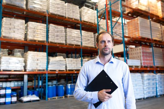 Front view of handsome mature Caucasian male supervisor holding clipboard and looking away in warehouse. This is a freight transportation and distribution warehouse. Industrial and industrial workers concept