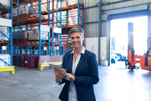 Front view of beautiful mature Caucasian female manager looking at camera while using digital tablet in warehouse. This is a freight transportation and distribution warehouse. Industrial and industrial workers concept