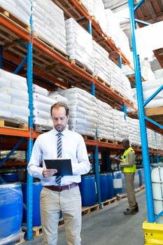 Front view of handsome mature Caucasian male supervisor checking stocks on clipboard in warehouse. African-american colleague is working in the background. This is a freight transportation and distribution warehouse. Industrial and industrial workers concept