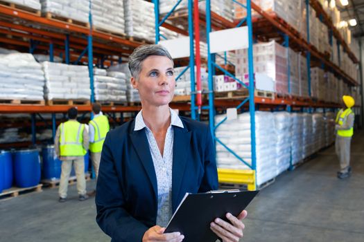 Front view of confident mature Caucasian female manager checking stocks on clipboard in warehouse. Diverse colleagues working in the background. This is a freight transportation and distribution warehouse. Industrial and industrial workers concept