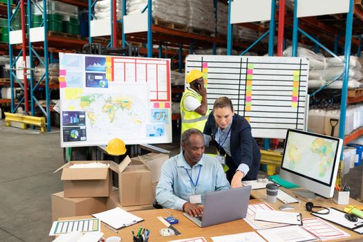 Front view of mature Caucasian female manager and African-american male supervisor discussing over laptop at desk in warehouse. This is a freight transportation and distribution warehouse. Industrial and industrial workers concept