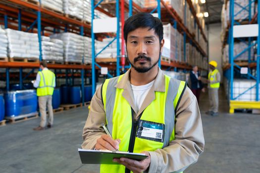 Close-up of handsome Asian male worker looking at camera while writing on clipboard in warehouse. This is a freight transportation and distribution warehouse. Industrial and industrial workers concept