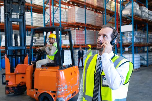 Front view of handsome Caucasian male supervisor talking on headset in warehouse. Diverse colleagues working in the background. This is a freight transportation and distribution warehouse. Industrial and industrial workers concept
