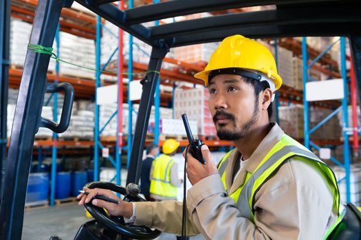 Portrait close-up of mature Caucasian male worker talking on walkie-talkie while driving forklift in warehouse. This is a freight transportation and distribution warehouse. Industrial and industrial workers concept