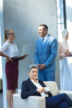 Front view of mature businesswoman drinking coffee and looking away in modern office. Behind her, colleagues interacting with each other.