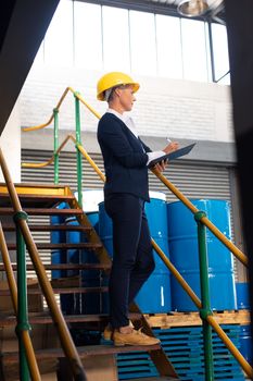 Low angle view of Caucasian female manager writing on clipboard on stairs in warehouse. This is a freight transportation and distribution warehouse. Industrial and industrial workers concept