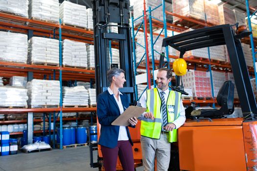 Front view of happy mature Caucasian male and female staff interacting with each other near forklift in warehouse. This is a freight transportation and distribution warehouse. Industrial and industrial workers concept