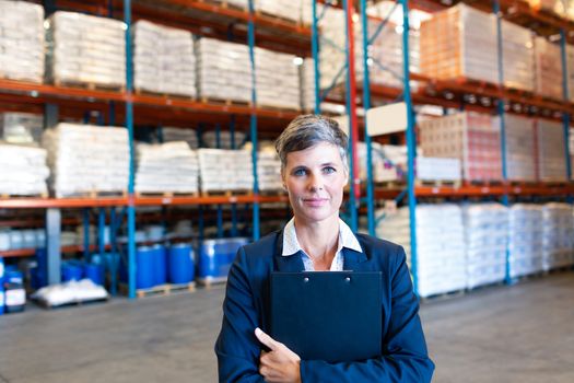 Portrait of beautiful mature Caucasian female manager holding clipboard and looking at camera in warehouse. This is a freight transportation and distribution warehouse. Industrial and industrial workers concept