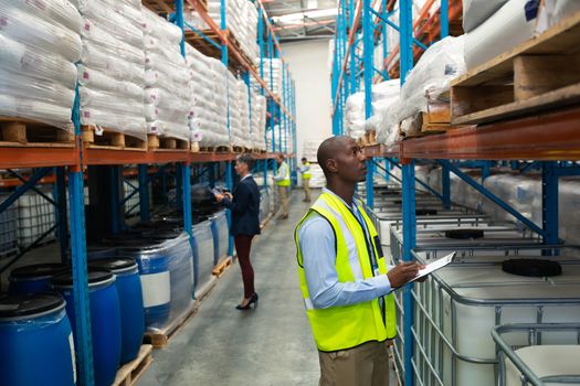 Side view of diverse warehouse staff checking stocks in aisle in warehouse. They are holding clipboards and writing in it. This is a freight transportation and distribution warehouse. Industrial and industrial workers concept