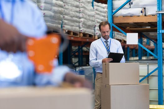 Front view of handsome mature Caucasian male supervisor working on clipboard while African-american staff works on the foreground in warehouse. This is a freight transportation and distribution warehouse. Industrial and industrial workers concept