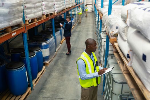 High angle side view of diverse warehouse staff checking stocks in aisle in warehouse. They are holding clipboards and writing in it. This is a freight transportation and distribution warehouse. Industrial and industrial workers concept