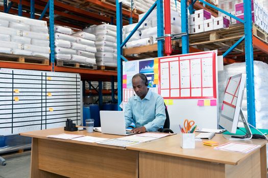 Front view of mature African-american male supervisor working on laptop at desk in warehouse. This is a freight transportation and distribution warehouse. Industrial and industrial workers concept
