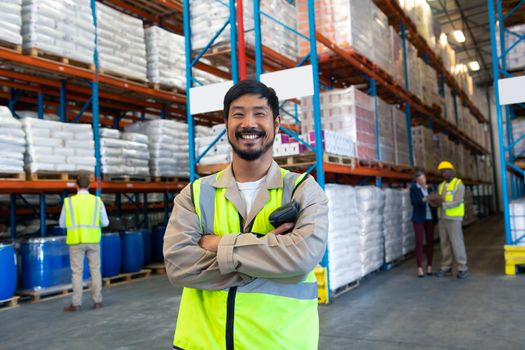 Front view of handsome Asian male worker standing with arms crossed and looking at camera in warehouse. Diverse colleagues working in the background. This is a freight transportation and distribution warehouse. Industrial and industrial workers concept