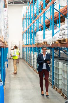 Front view of pretty Caucasian female manager working on clipboard while walking in warehouse. Diverse male staff work behind her. This is a freight transportation and distribution warehouse. Industrial and industrial workers concept