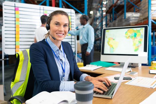 Front view of mature Caucasian female manager with headset using laptop at desk in warehouse. In the background diverse coworkers are discussing in front of whiteboard. This is a freight transportation and distribution warehouse. Industrial and industrial workers concept