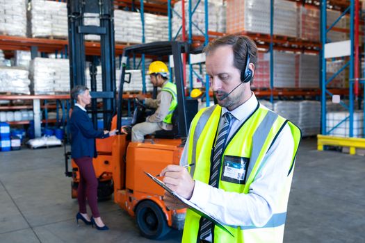 Front view of handsome Caucasian male supervisor with headset writing on clipboard in warehouse. Diverse colleagues working in the background. This is a freight transportation and distribution warehouse. Industrial and industrial workers concept
