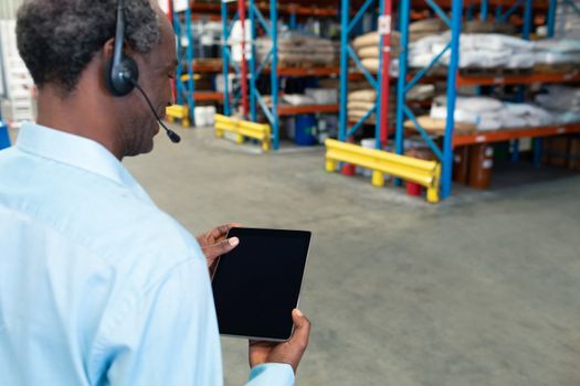 Rear view of mature African american male supervisor with headset using digital tablet in warehouse. This is a freight transportation and distribution warehouse. Industrial and industrial workers concept