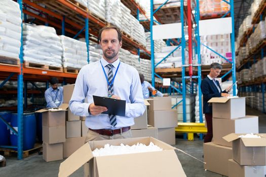 Front view of handsome Caucasian male supervisor checking stocks and looking at camera in warehouse. Diverse warehouse workers unpacking cardboard boxes. This is a freight transportation and distribution warehouse. Industrial and industrial workers concept