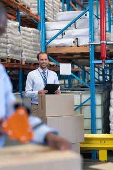 Front view of happy Caucasian male supervisor with clipboard looking at camera in warehouse. African-american staff works on the foreground. This is a freight transportation and distribution warehouse. Industrial and industrial workers concept