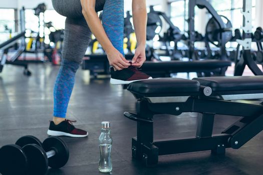 Low section of young Caucasian female athletic tying shoe laces in fitness center. Bright modern gym with fit healthy people working out and training