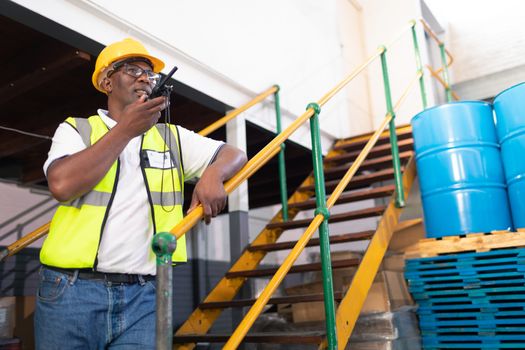 Low angle view of African-american male worker talking on talkie walkie in warehouse. This is a freight transportation and distribution warehouse. Industrial and industrial workers concept